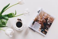 Top view of a coffee cup, flower vase, and magazine on a white table, perfect for relaxation.